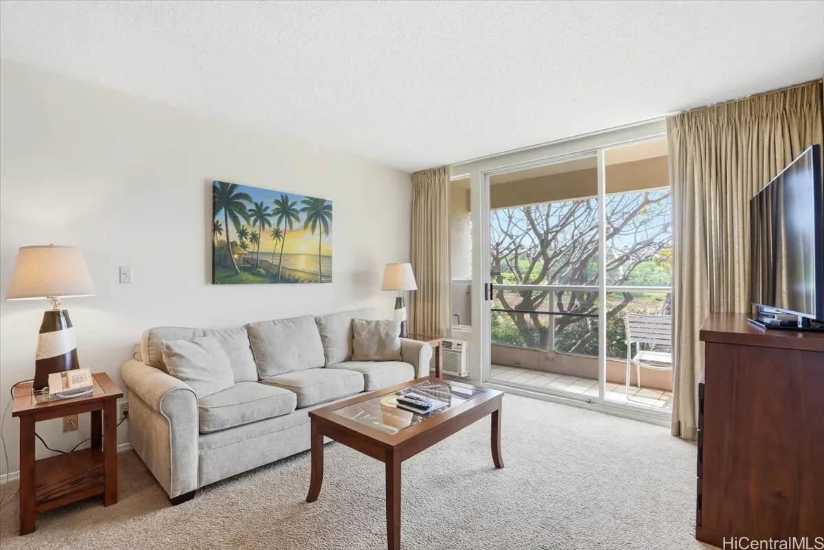 Living room with beige sofa, wood table, and palm tree painting. Balcony with trees visible.