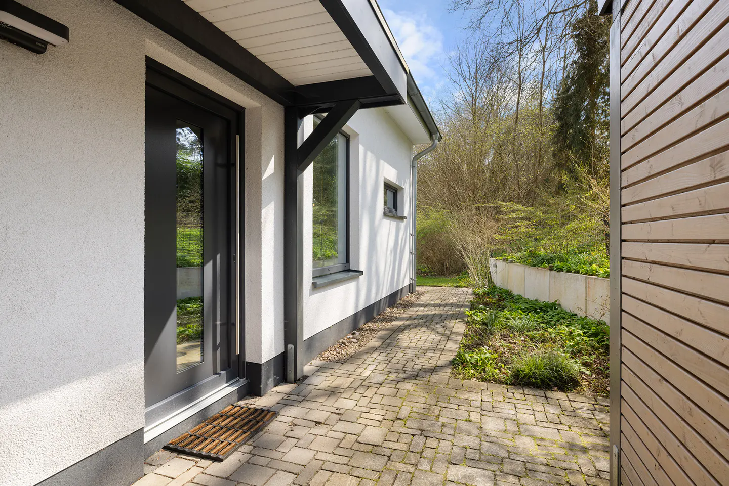 Exterior view of a modern white house with black trim, a brick walkway, and a wooden wall on the right.