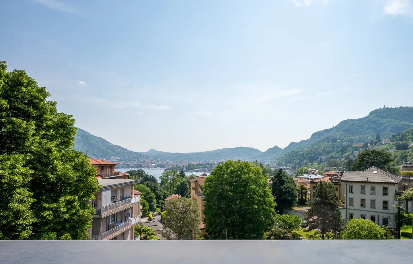 Scenic view of Lake Como, Italy, with mountains, buildings, and lush green trees under a clear blue sky.