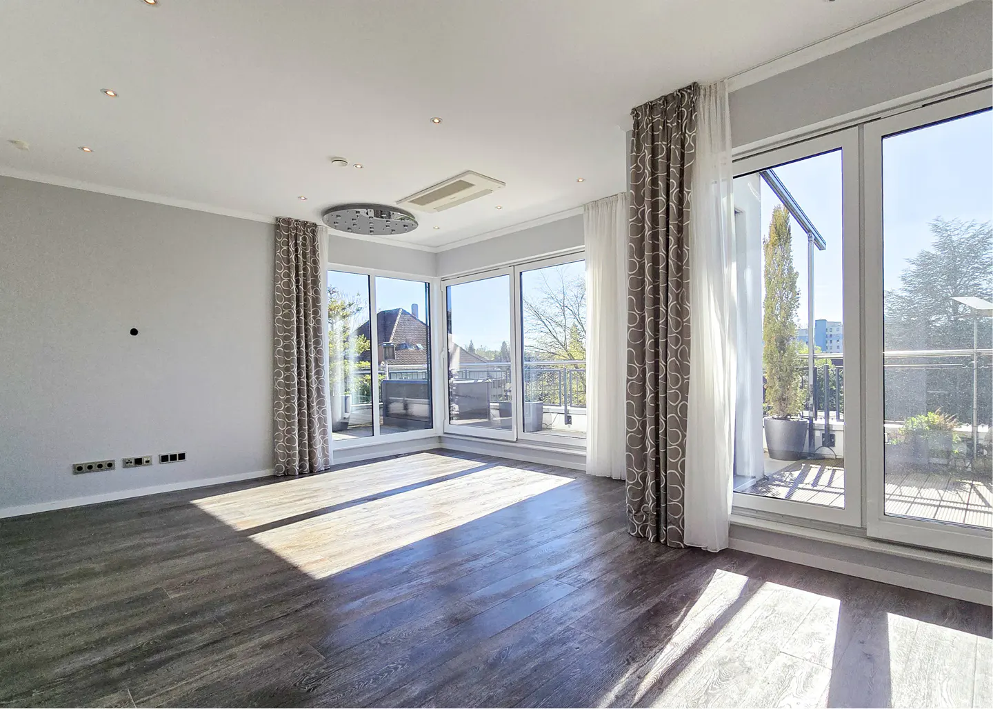 Bright, empty room with gray walls, dark wood floors, and large windows with patterned curtains leading to a balcony.