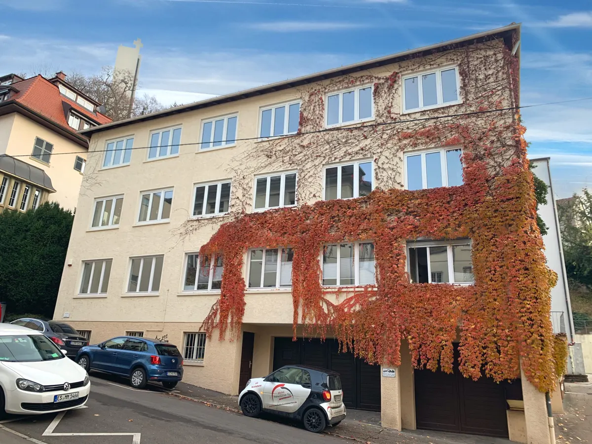 Three-story apartment building with beige facade and red ivy. Cars parked on the street. Blue sky.