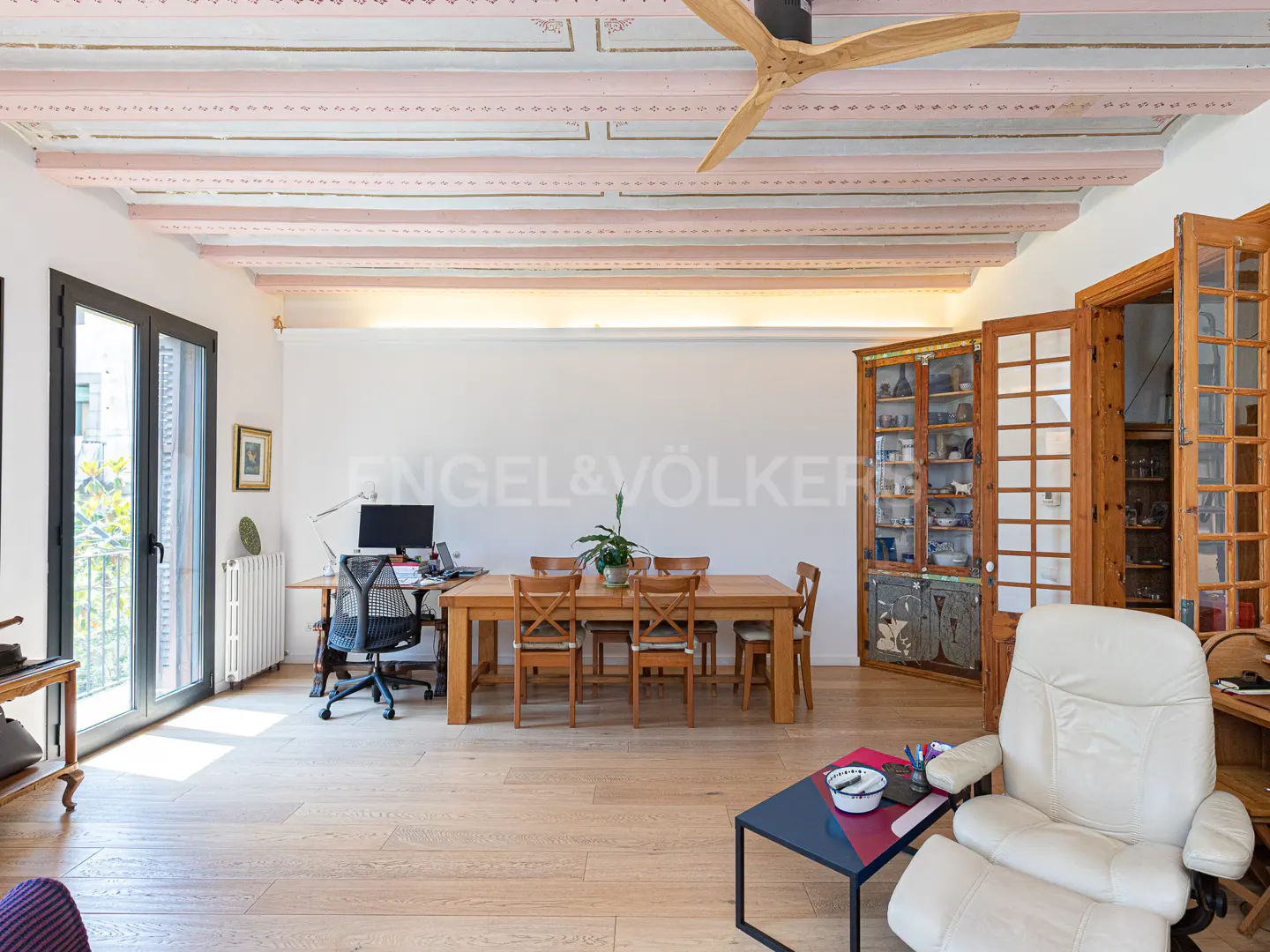Bright living room with wood floors, a dining table, desk, and white leather chair. The ceiling has decorative beams and a modern fan.