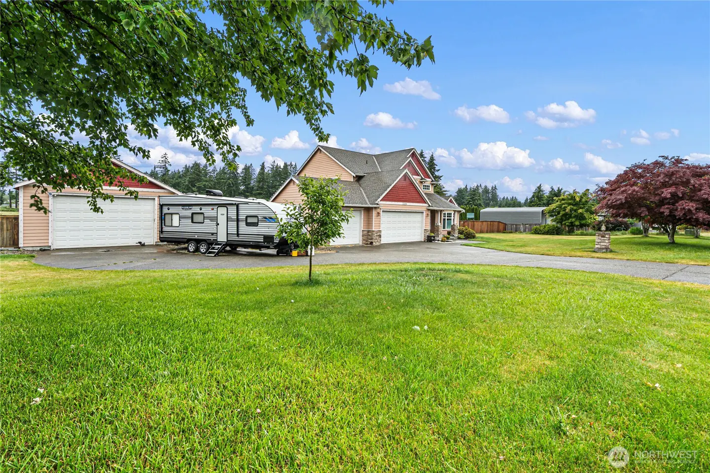 A tan and red house with a large garage and a parked travel trailer on a spacious green lawn under a blue sky.
