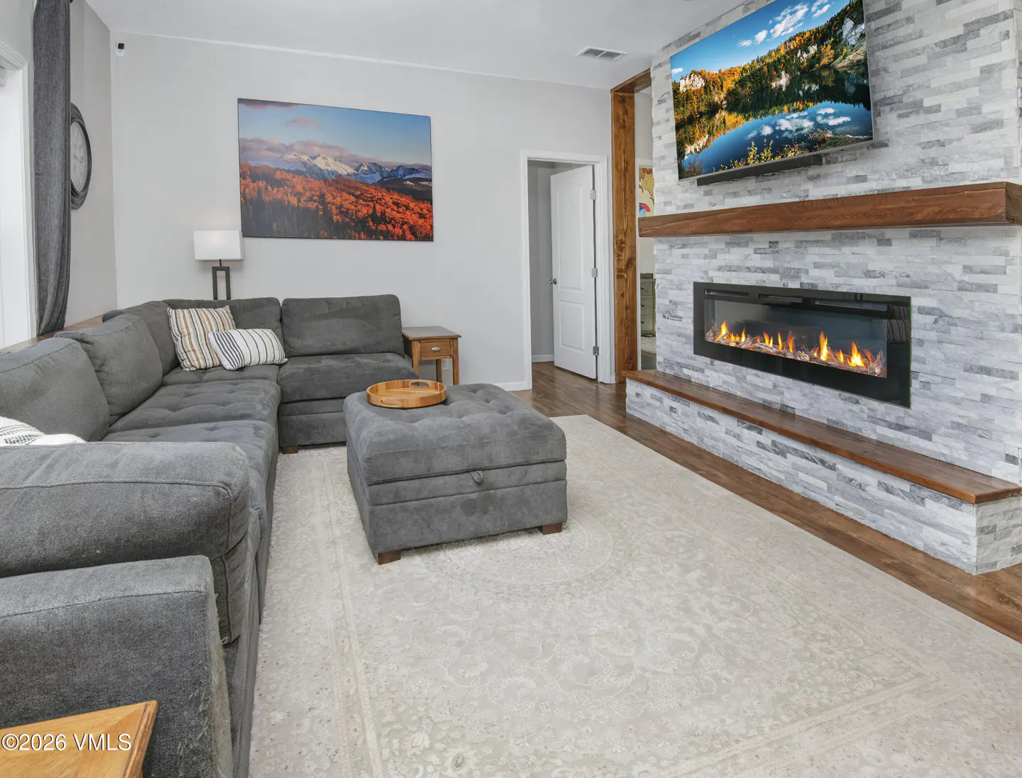 Living room with gray sectional sofa, ottoman, and stone fireplace with TV above. Artwork on the wall.