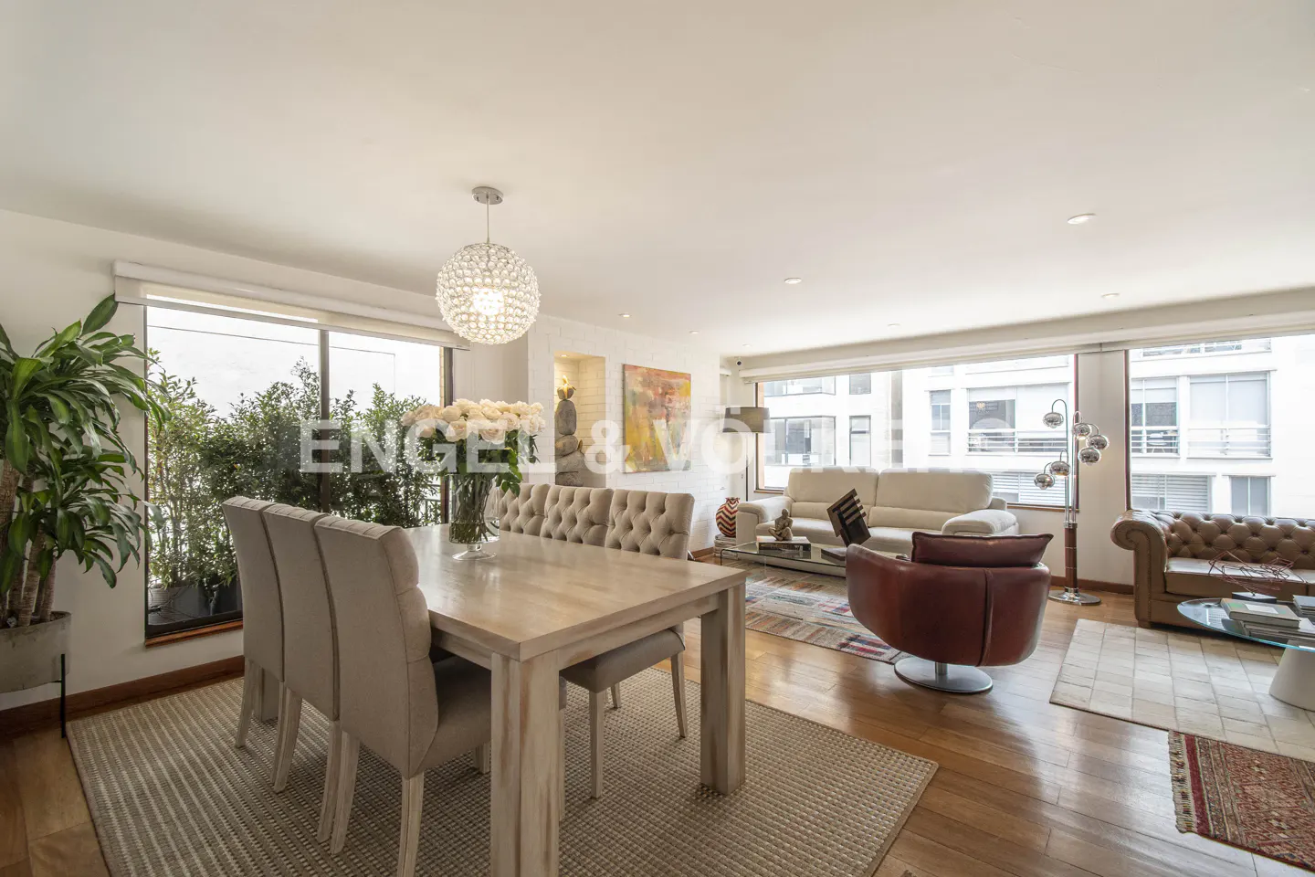 Bright open-plan dining and living room with light wood floors, beige tufted chairs, and large windows overlooking trees.
