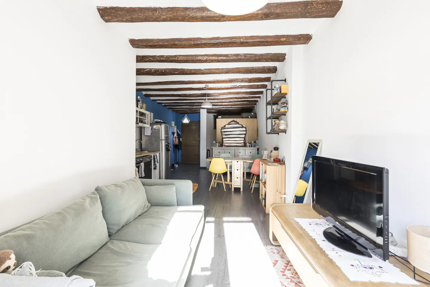 Bright, narrow living room with exposed wood beams, a light green sofa, and a TV. The kitchen is visible in the background.