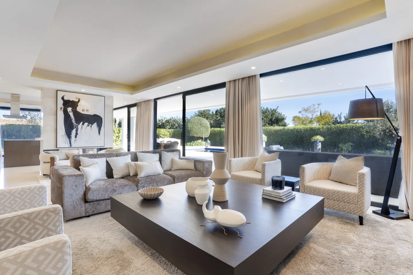 Bright living room with a gray sofa, white pillows, a large dark wood coffee table, and floor-to-ceiling windows.