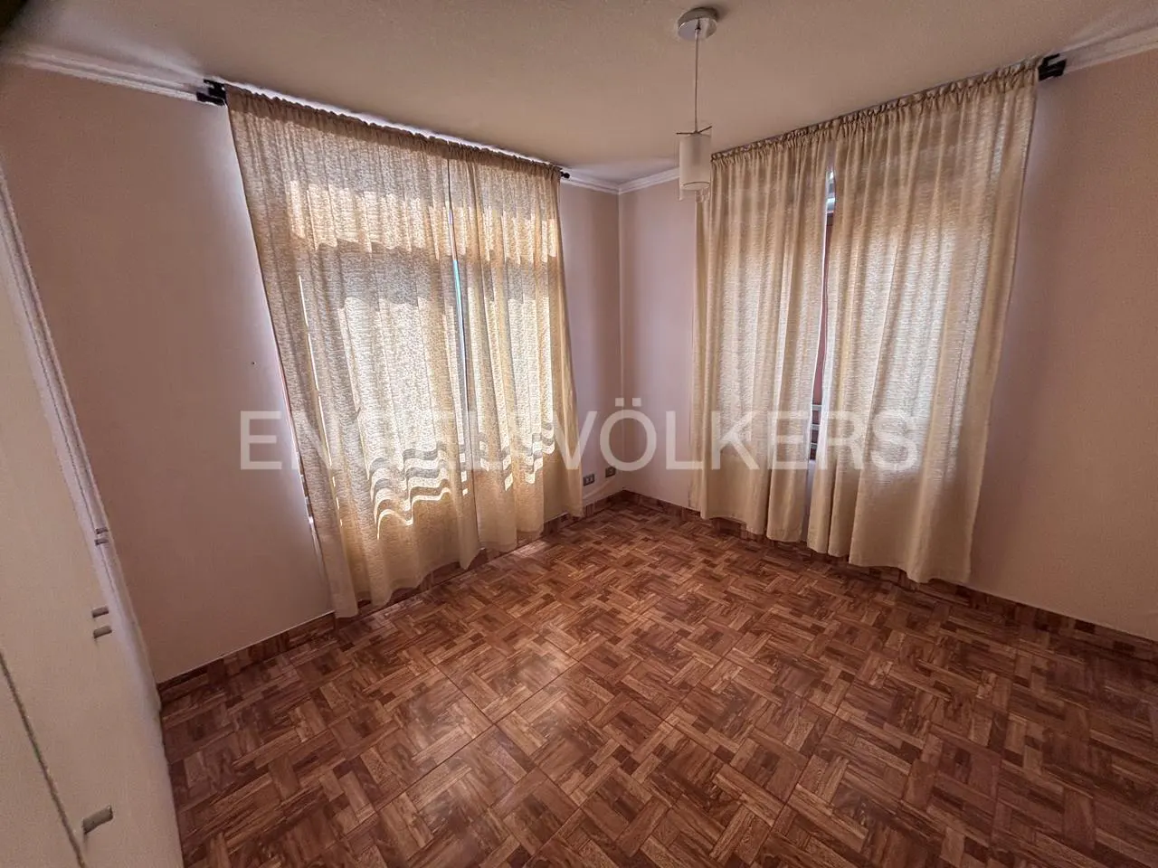 Empty bedroom with pink walls, brown parquet floor, and beige curtains covering two windows. A white pendant light hangs from the ceiling.