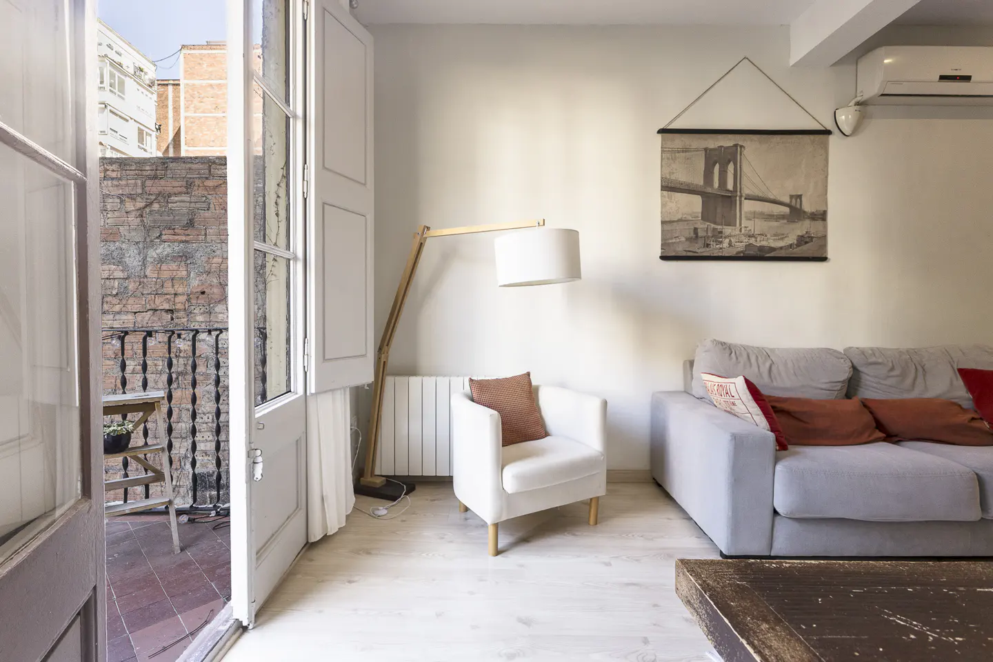Living room with a white armchair, grey sofa, floor lamp, and a balcony door opening to a brick-walled terrace.