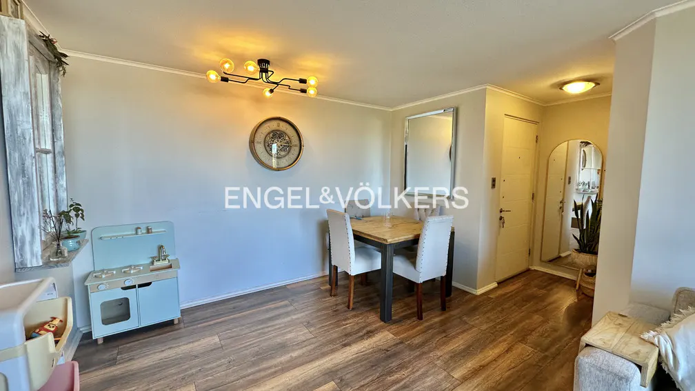 A living room with wood floors, a dining table with two chairs, and a child's play kitchen. A clock hangs on the wall.