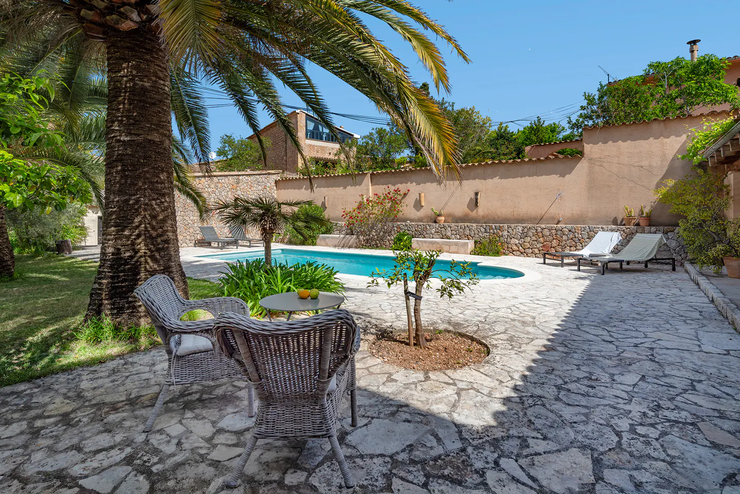 Outdoor patio with stone flooring, pool, and palm trees. Wicker chairs and a small table are in the foreground.
