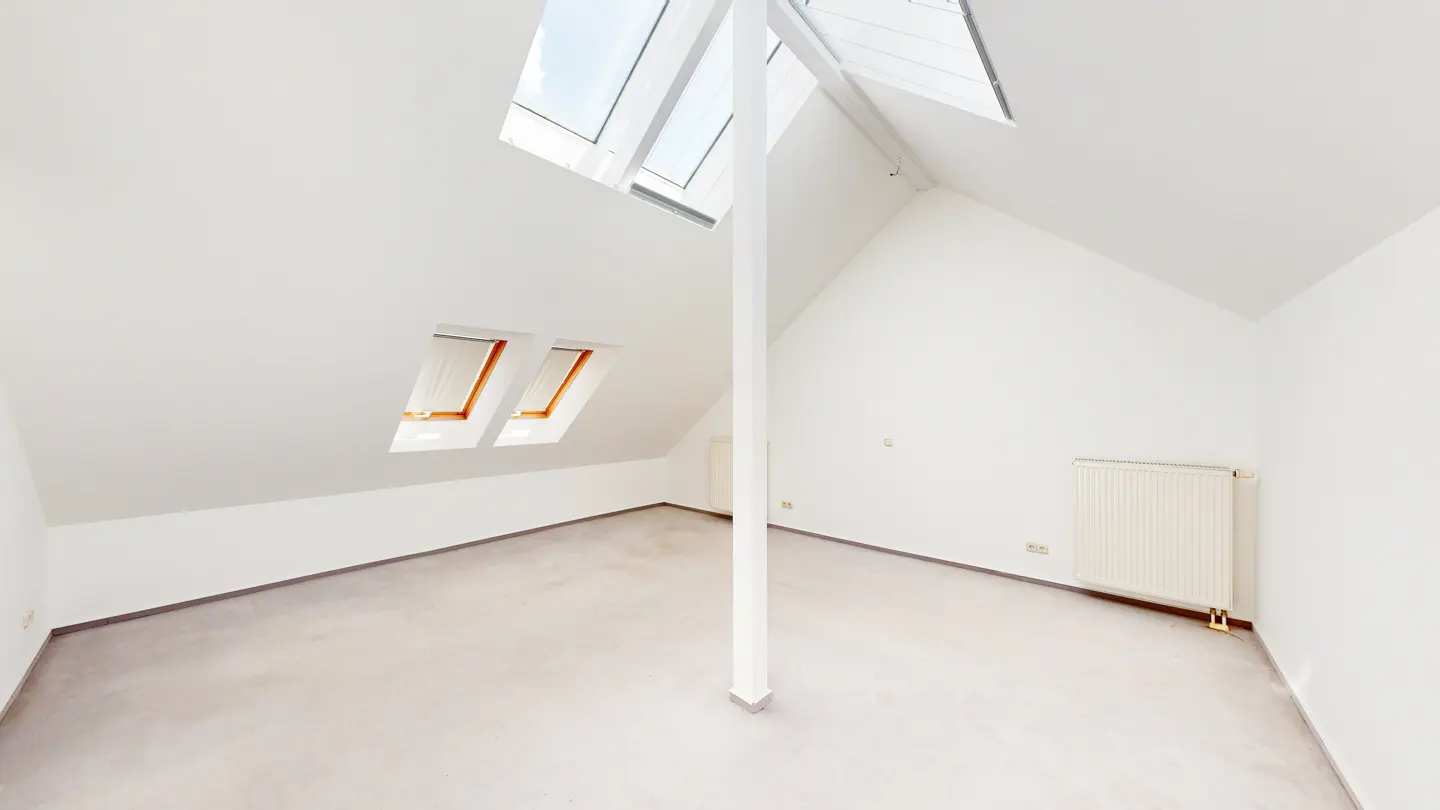 Bright, empty attic room with white walls, gray floor, skylights, and a support beam. A radiator is on the right wall.