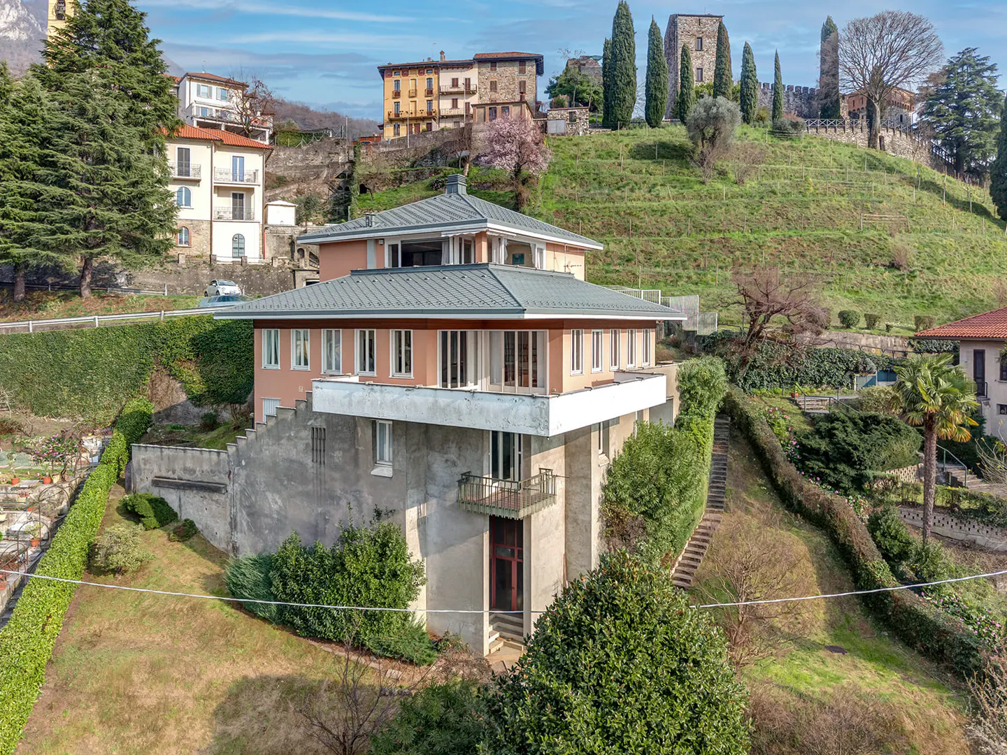 Exterior view of a two-story peach house with a gray roof, surrounded by greenery and other buildings on a hillside.