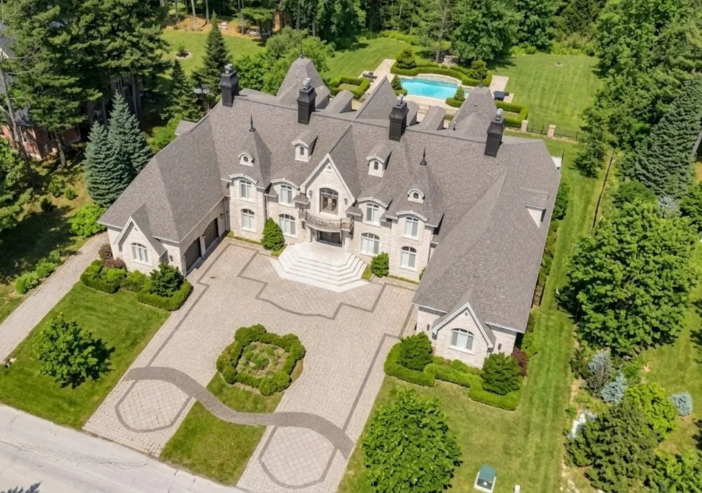 Aerial view of a large, light-stone mansion with a gray roof, black chimneys, and a paved driveway with manicured landscaping. A pool is visible in the background.