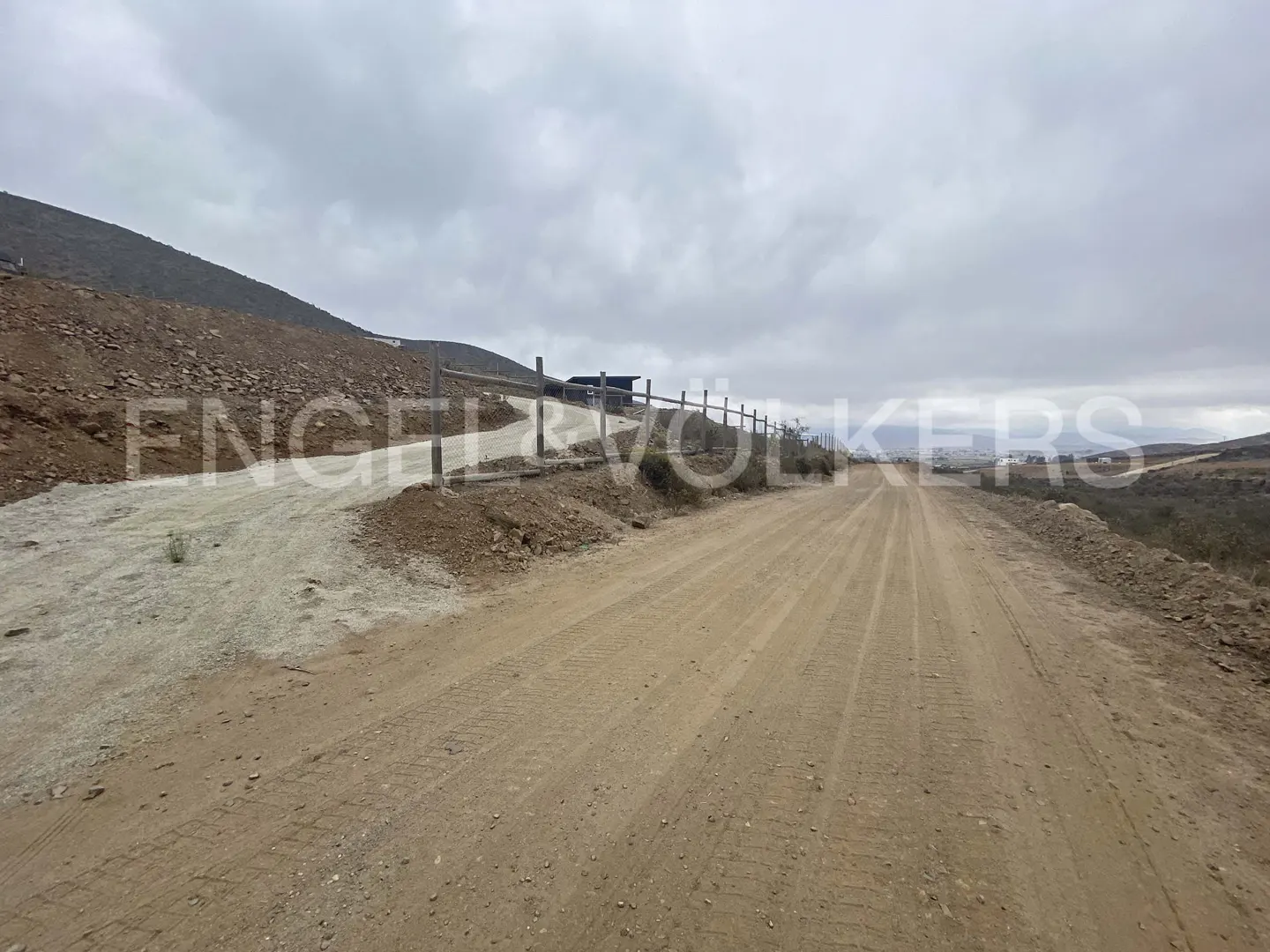 Dirt road leading uphill with a wooden fence on the left, under a cloudy sky.