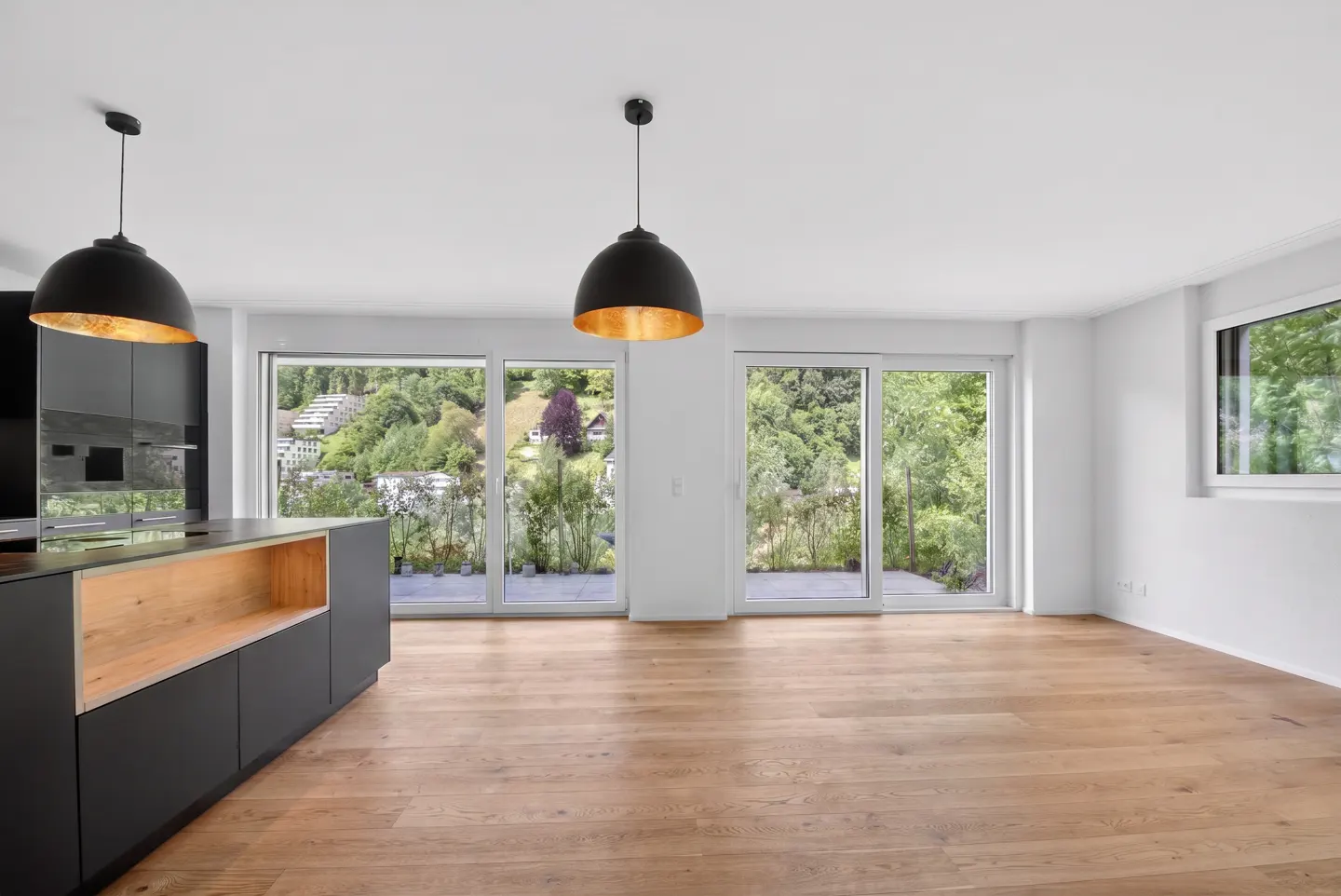 Bright, modern kitchen with wood floors, black cabinets, and black pendant lights. Large windows offer a view of lush green hills.