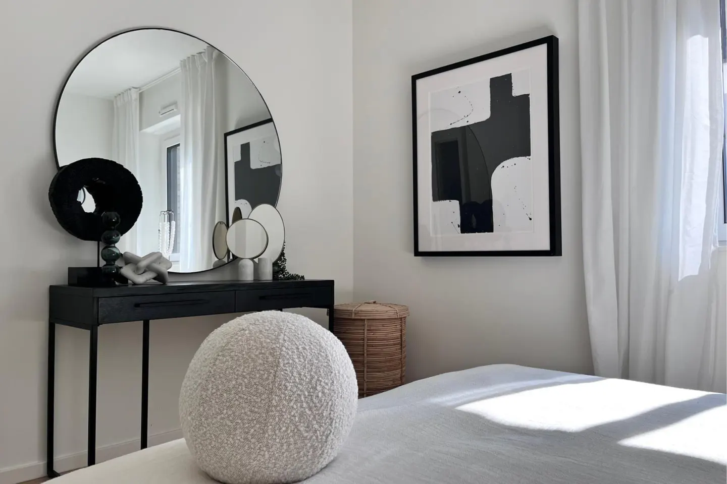 Bright bedroom with a black vanity, round mirror, and abstract art. A white, textured round pillow sits on the bed.