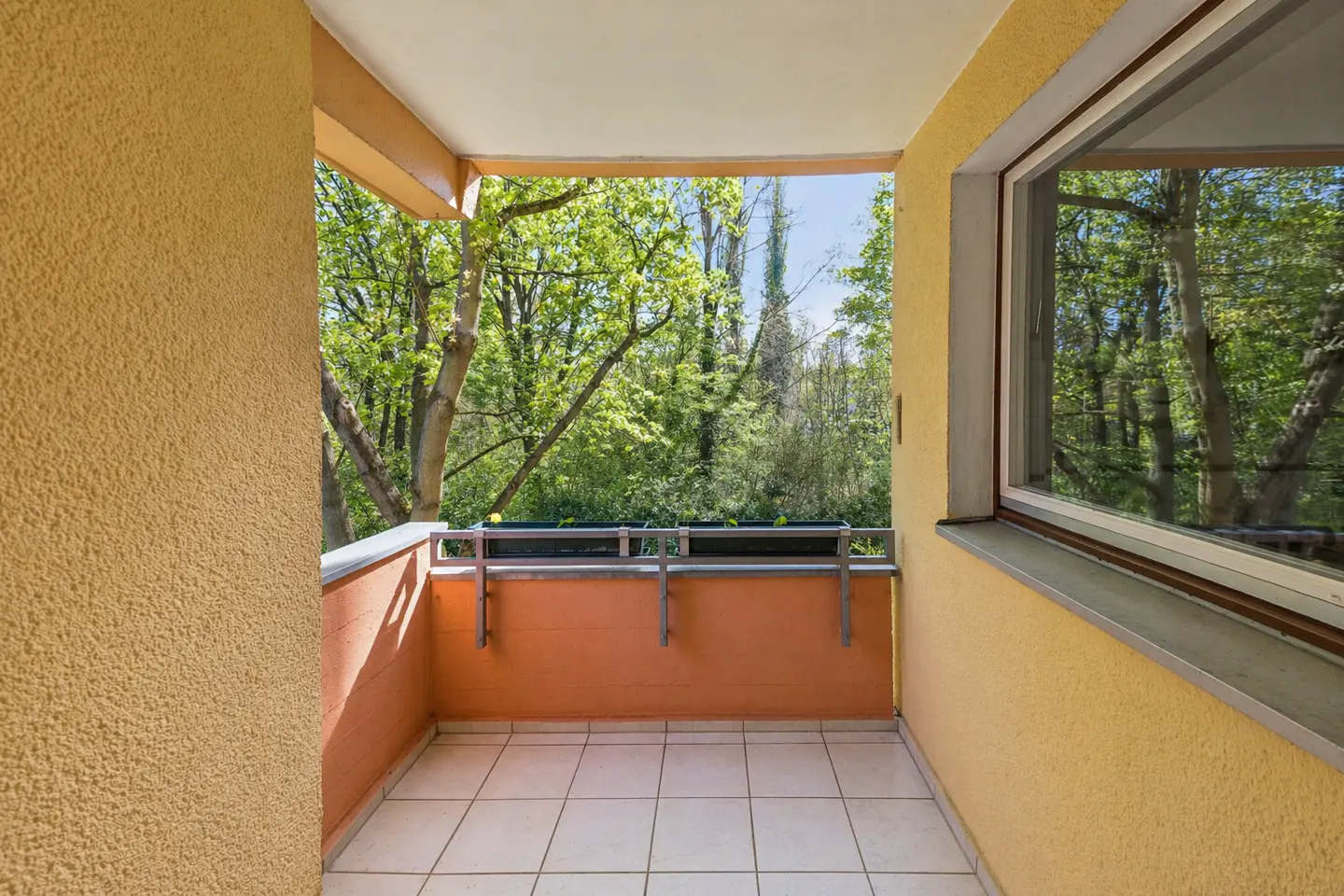 Balcony with yellow walls, white tiled floor, and orange railing overlooking green trees. A window reflects the trees.