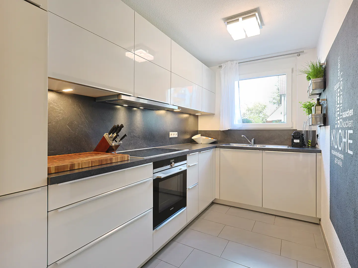 Bright kitchen with white cabinets, gray countertops, and gray tile floor. A window provides natural light. A chalkboard wall adds a decorative touch.