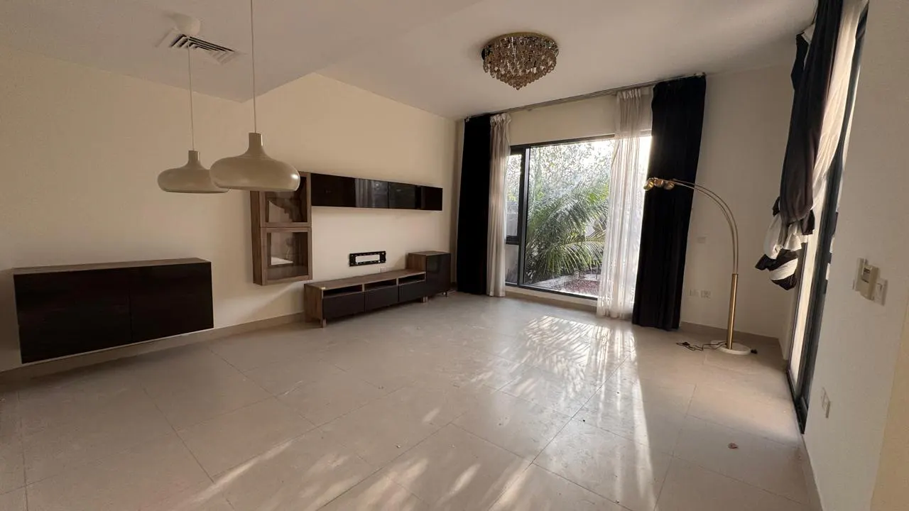 A bright, empty living room with tile floors, dark wood cabinets, and a large window with black and white curtains.