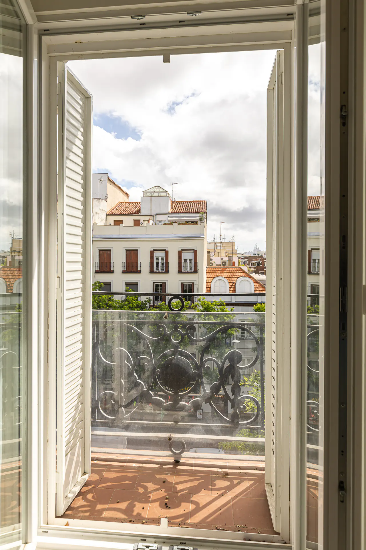 View from open white shuttered doors onto a balcony with wrought iron railing, overlooking city buildings under a cloudy sky.
