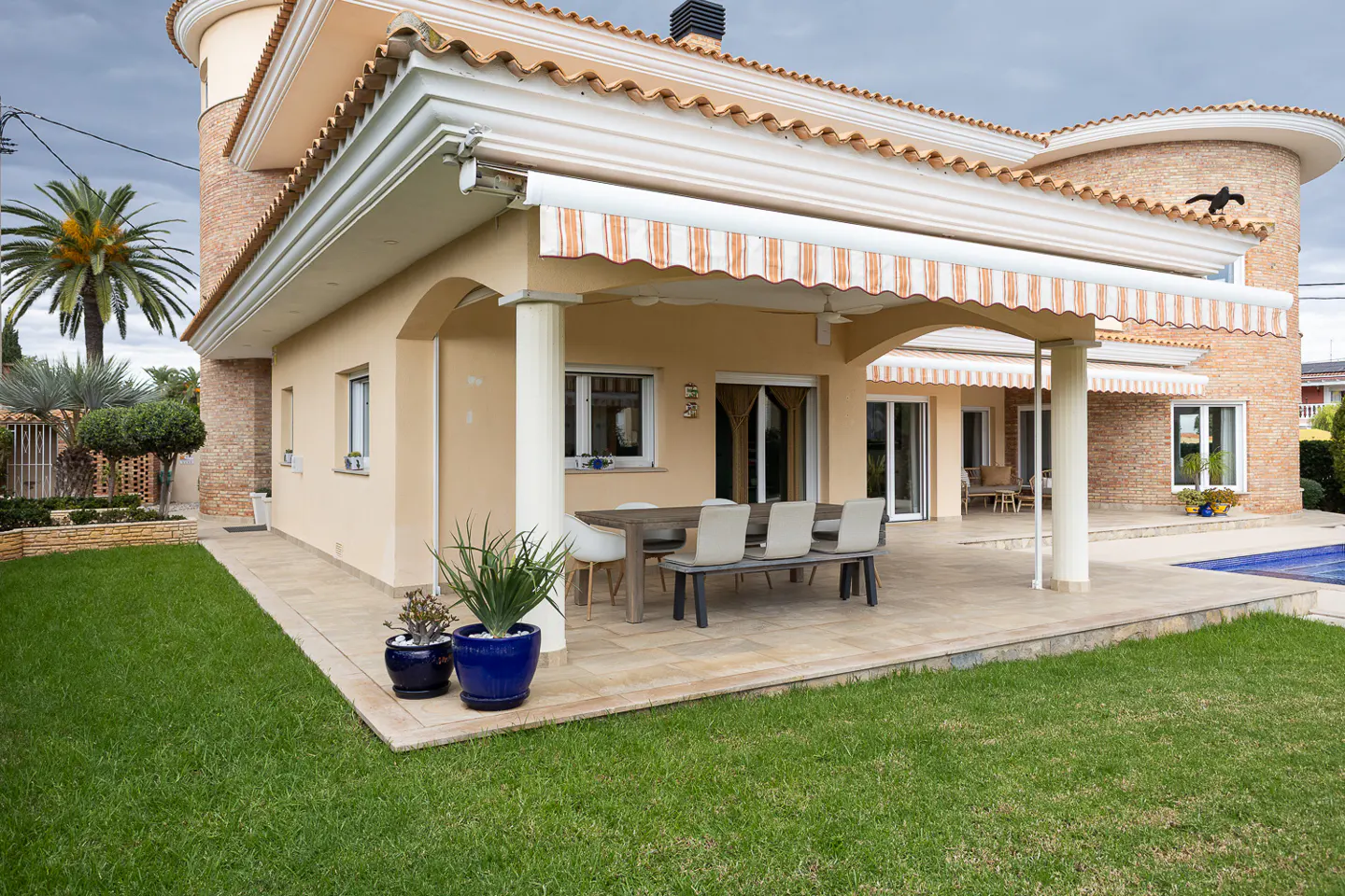 Beige house with a striped awning over a patio with a table and chairs, next to a green lawn.