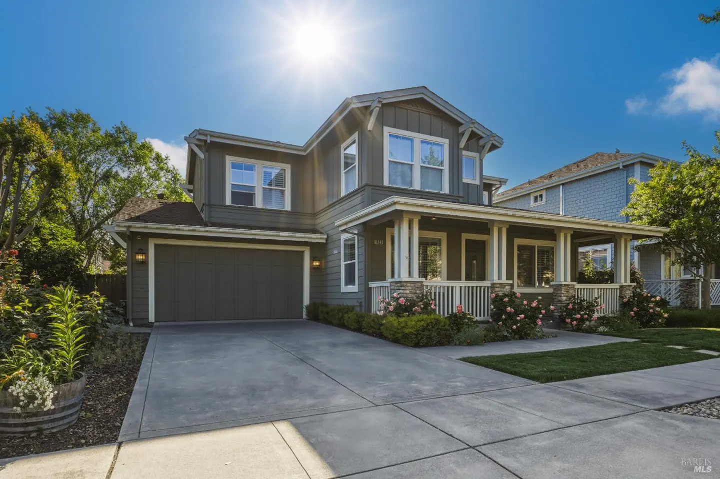 Two-story gray house with a front porch, white railings, and stone pillars under a bright blue sky.