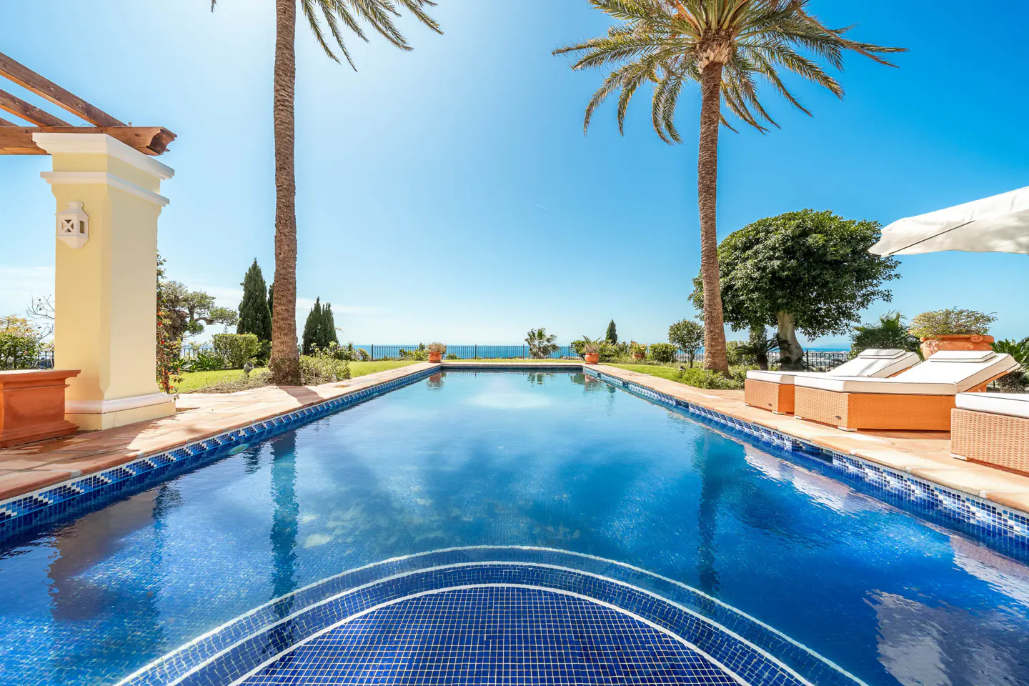 A luxurious blue tiled pool with palm trees, lounge chairs, and ocean view under a clear blue sky.