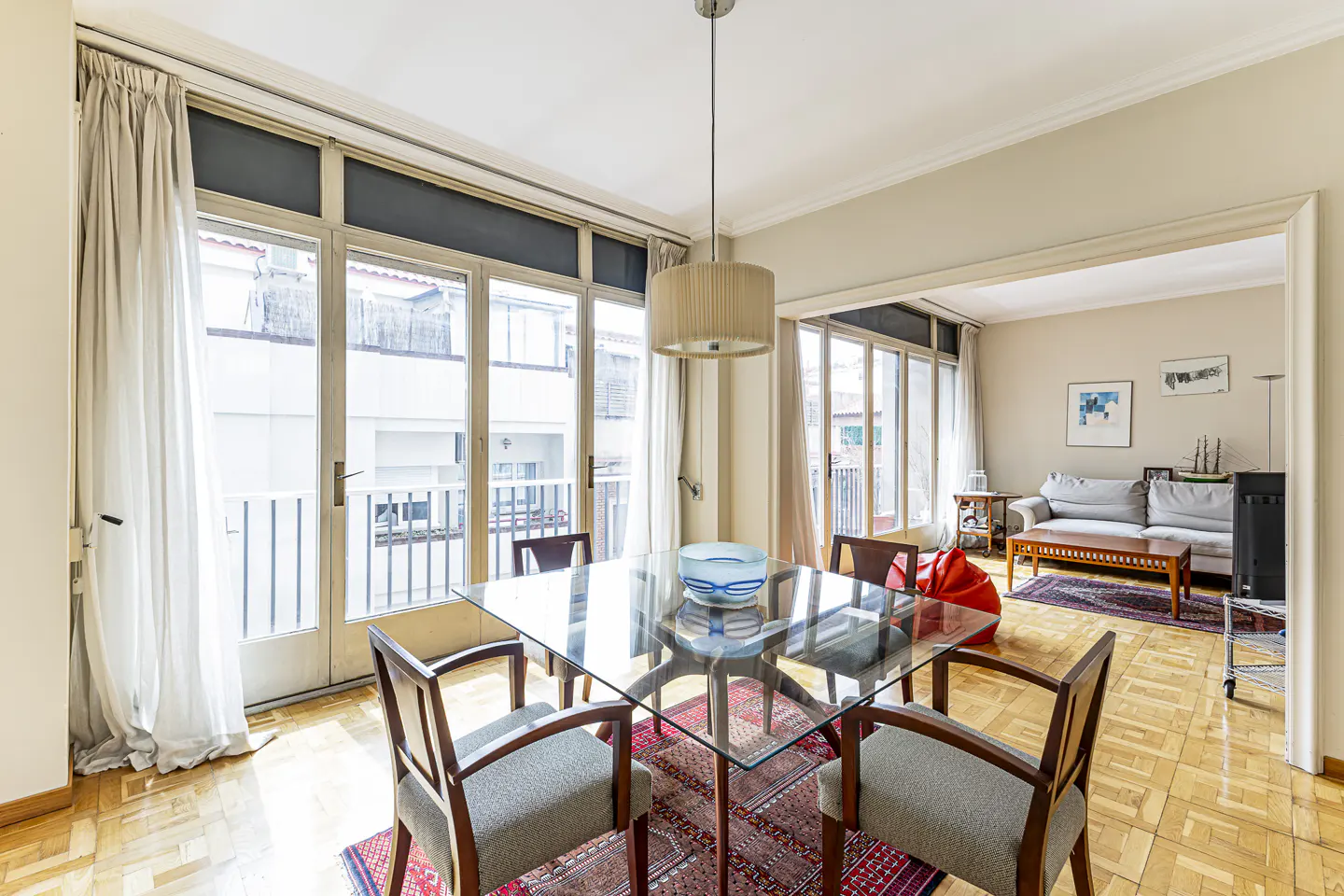 Dining area with a glass table, four chairs, and a pendant light, opening into a living room with a sofa and balcony.