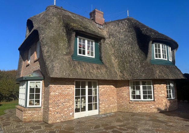 A two-story brick house with a thatched roof, white windows, and a stone patio on a sunny day.