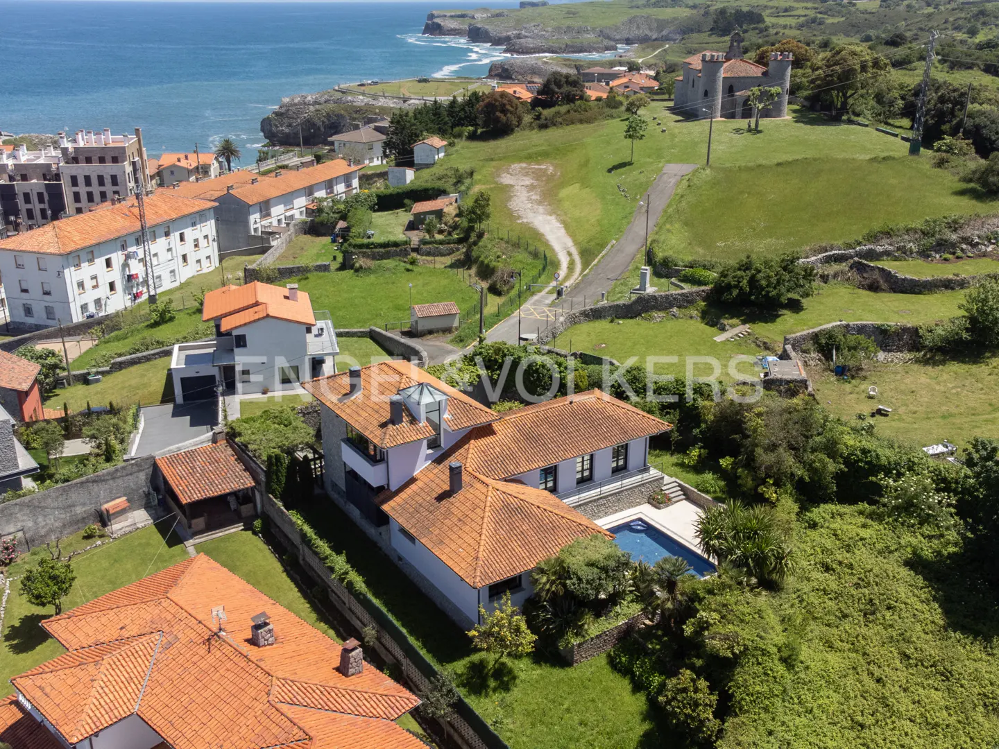 Aerial view of a white house with an orange tile roof and a blue pool, near the ocean and a castle.