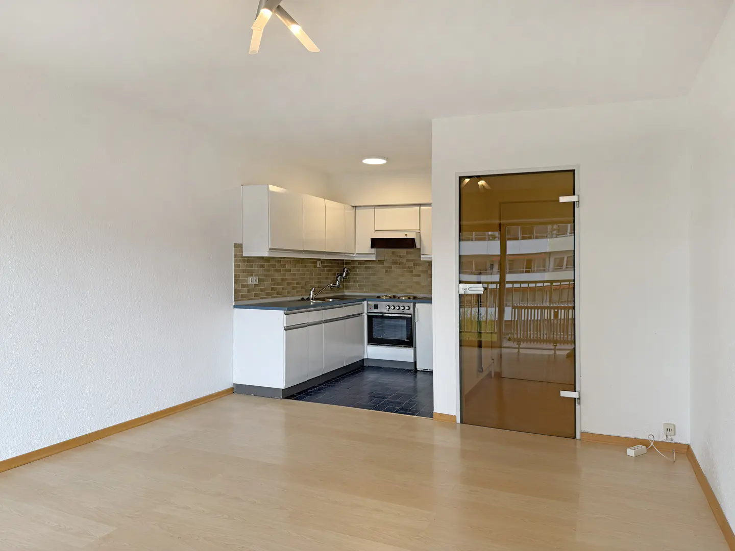 Bright, empty apartment with light wood floors, white walls, and a modern white kitchen with stainless steel appliances. A glass door reflects a building exterior.