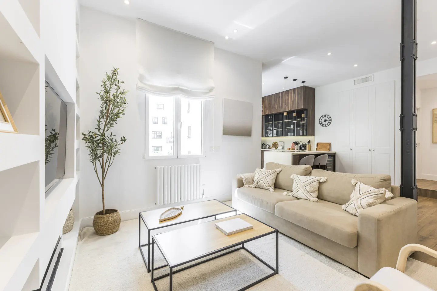 Bright living room with beige sofa, white walls, and black-framed coffee tables. A plant sits near a window with white blinds. The kitchen is visible in the background.