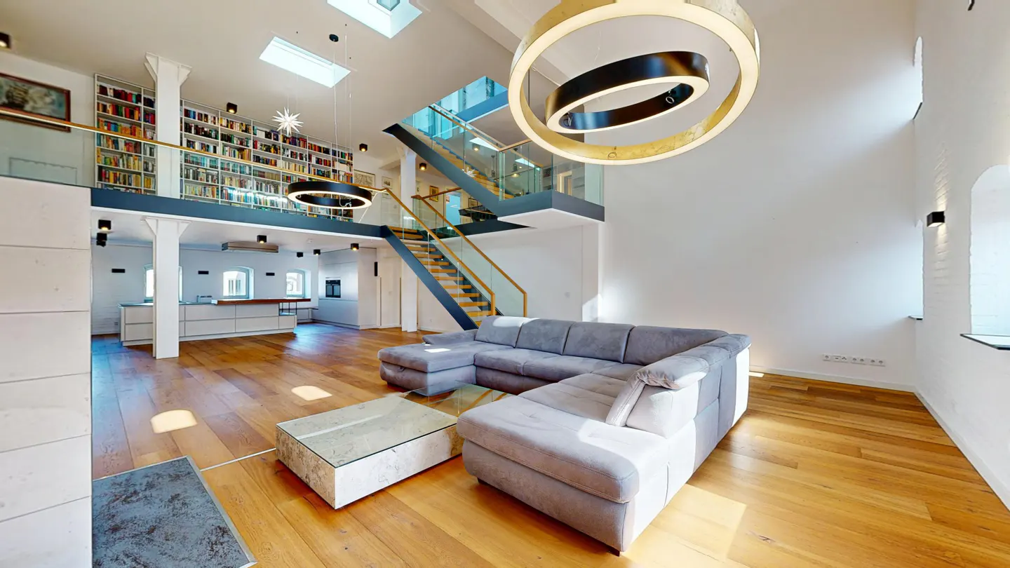 Bright, modern living room with hardwood floors, a gray sectional sofa, and a glass-topped coffee table. A staircase leads to a mezzanine with a full bookshelf.