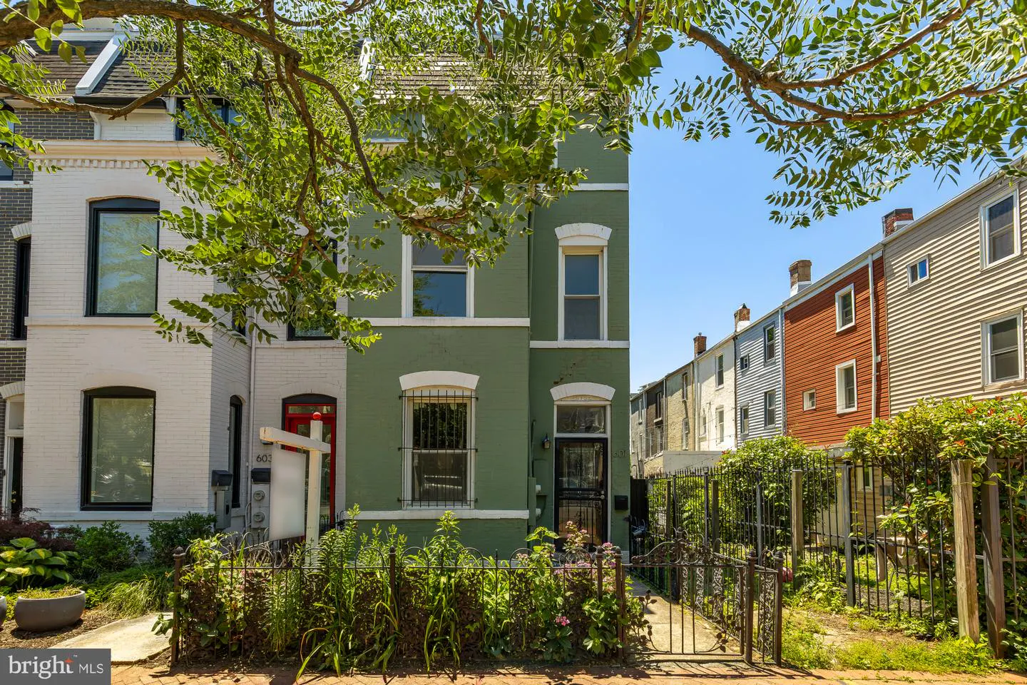 A green brick rowhouse with a red door and a for-sale sign, framed by tree branches under a clear blue sky.