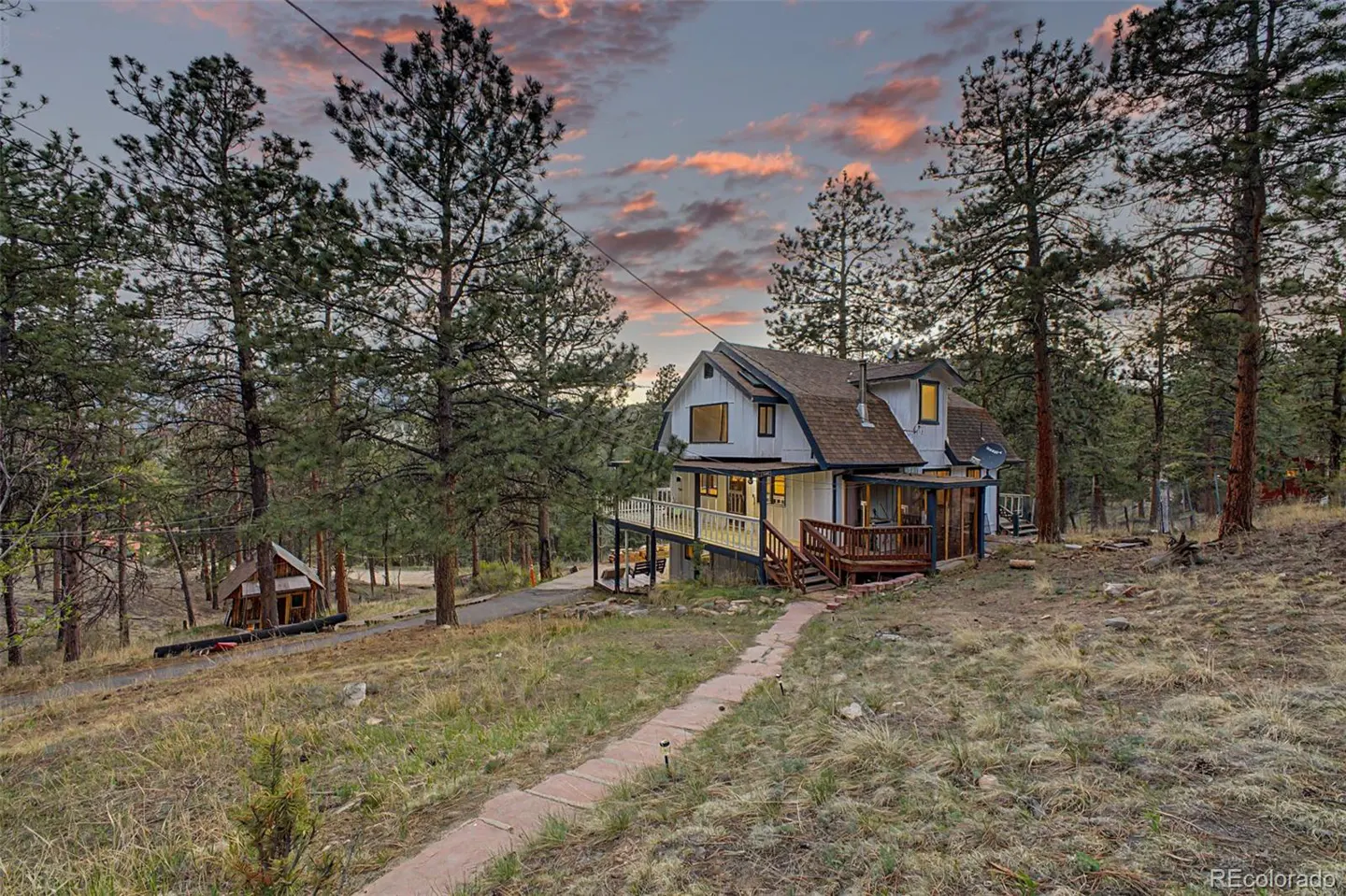 Exterior view of a white two-story house with a brown roof, surrounded by tall pine trees and a stone pathway leading to the entrance. The sky is a mix of blue and orange.