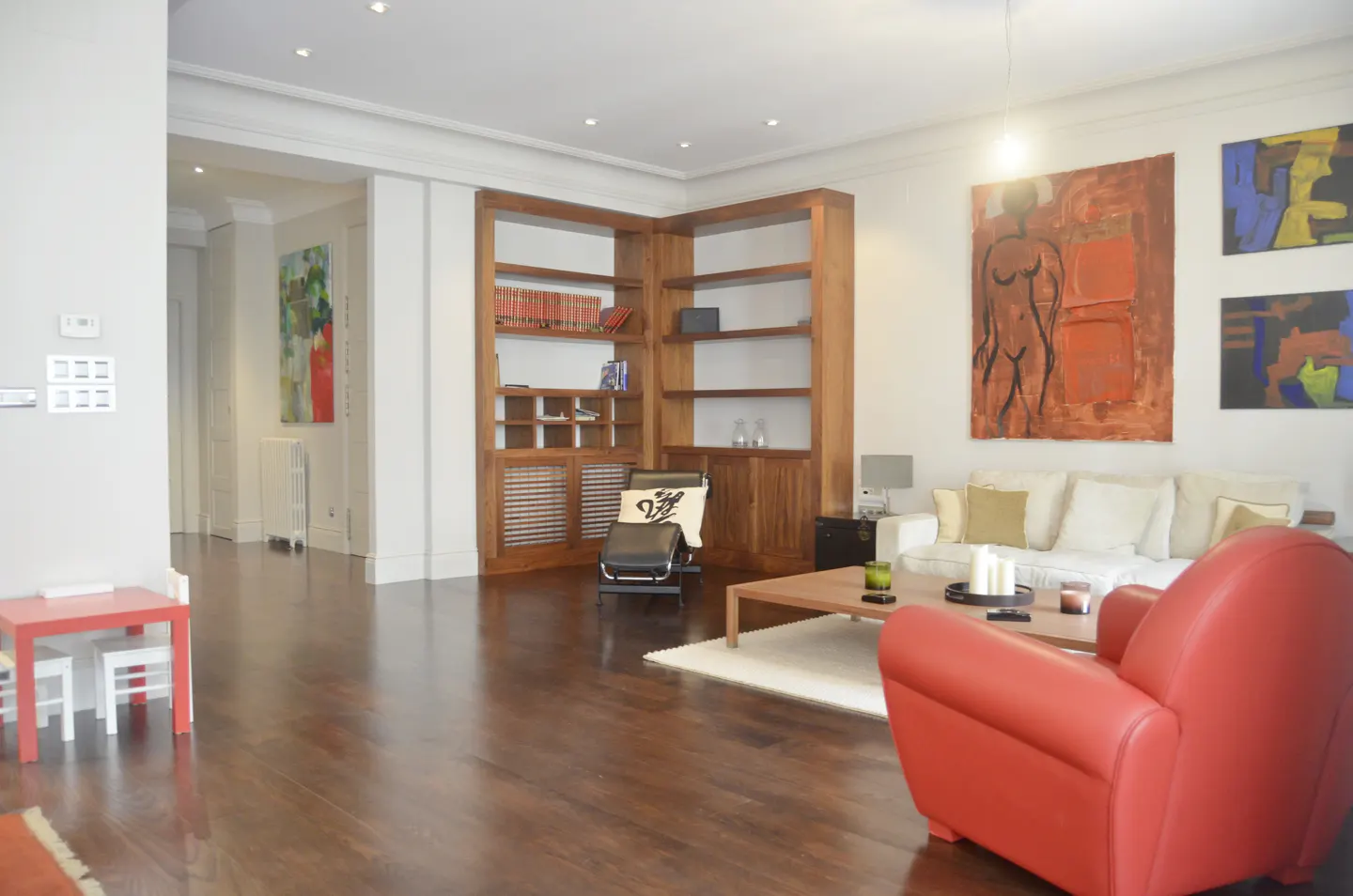 A living room with hardwood floors, a red armchair, a white sofa, and a wooden bookcase. Abstract art hangs on the wall.