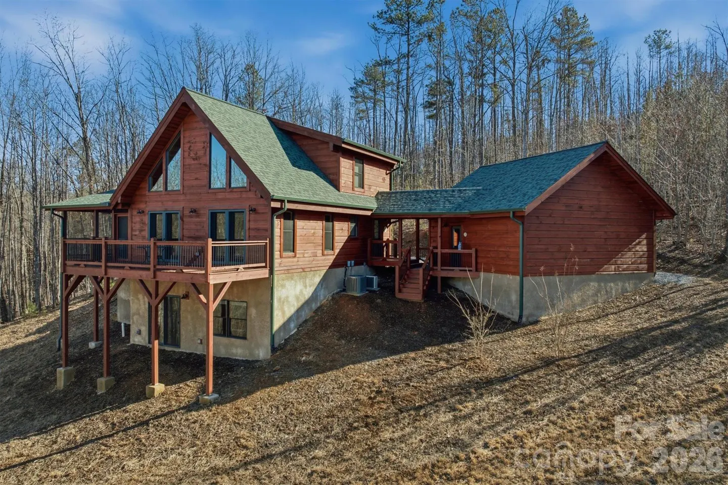 A two-story log cabin with a green roof sits on a grassy hill surrounded by bare trees. It has a deck and a walk-out basement.