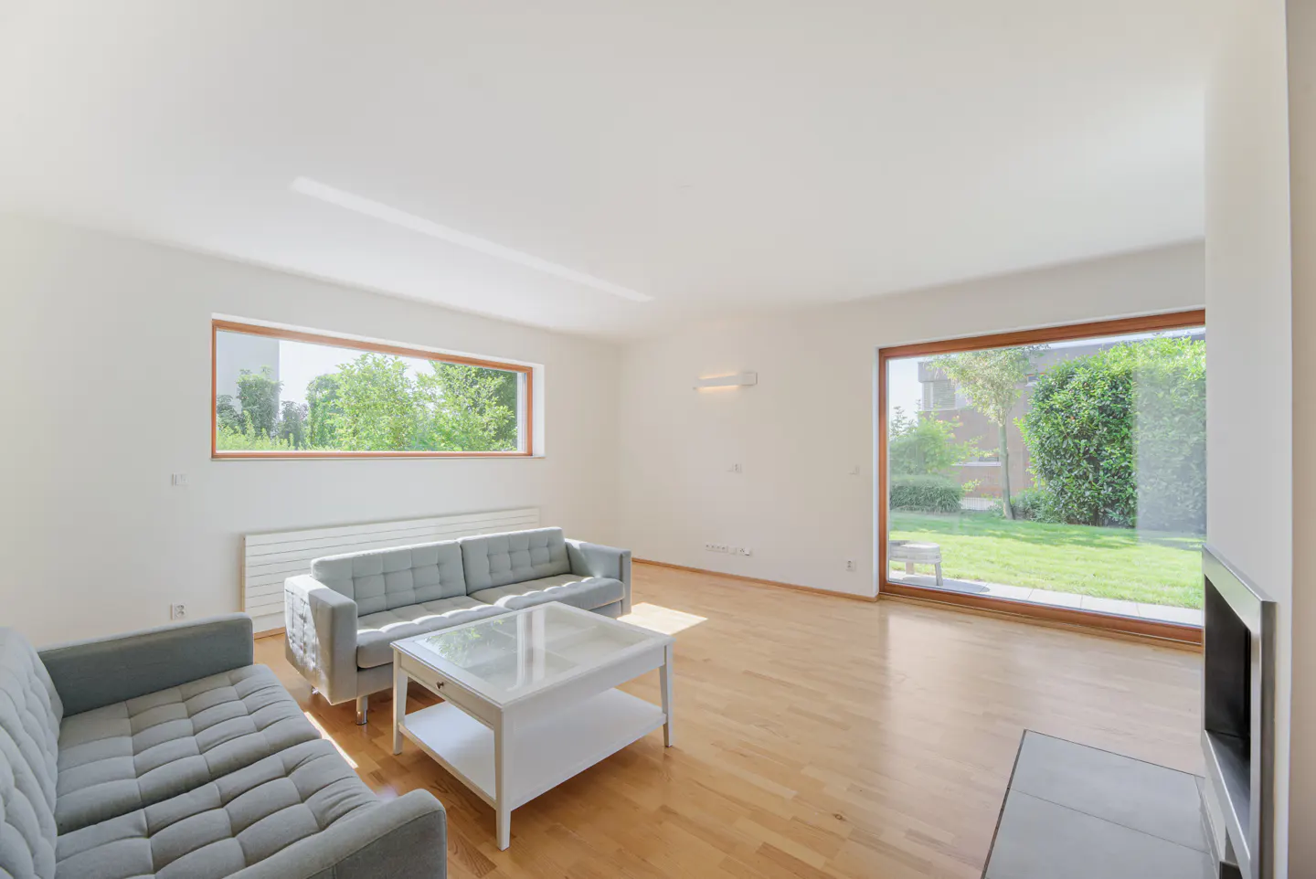 Bright living room with hardwood floors, gray sofas, and a white coffee table. Large windows offer views of a green yard.