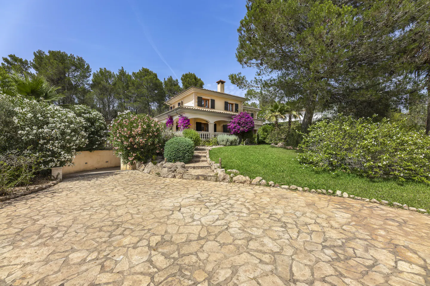 Two-story beige house with black shutters, stone driveway, green lawn, and colorful flowers under a blue sky.