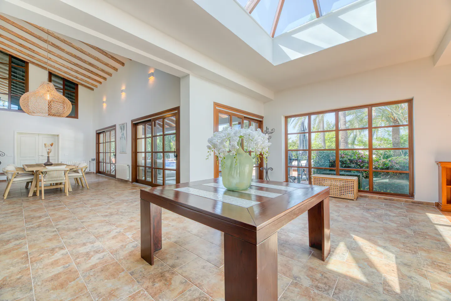 Bright, airy foyer with a dark wood table holding white flowers. Large windows and a skylight illuminate the tiled floor.