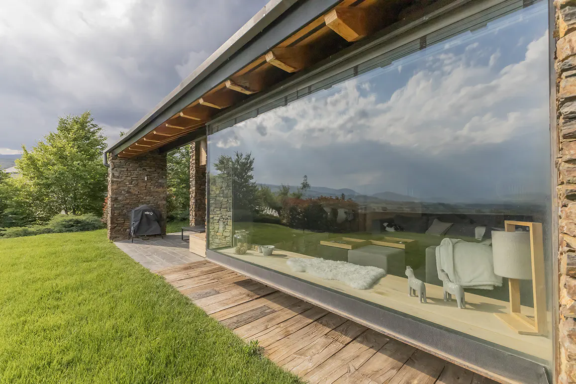 Modern home exterior with stone walls, wooden deck, and large floor-to-ceiling windows reflecting the cloudy sky.