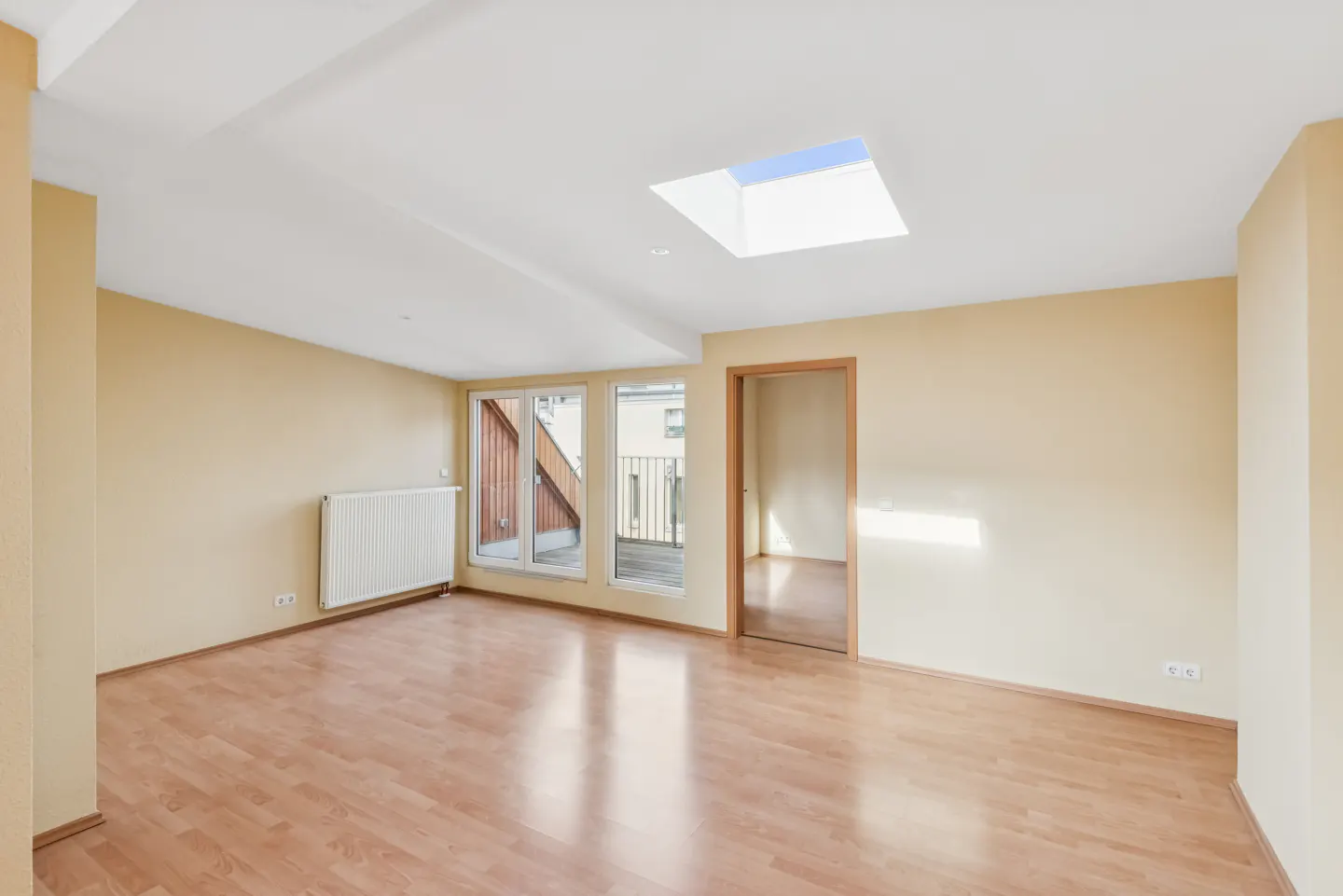Empty room with light-yellow walls, wood floor, radiator, skylight, and windows to a balcony.