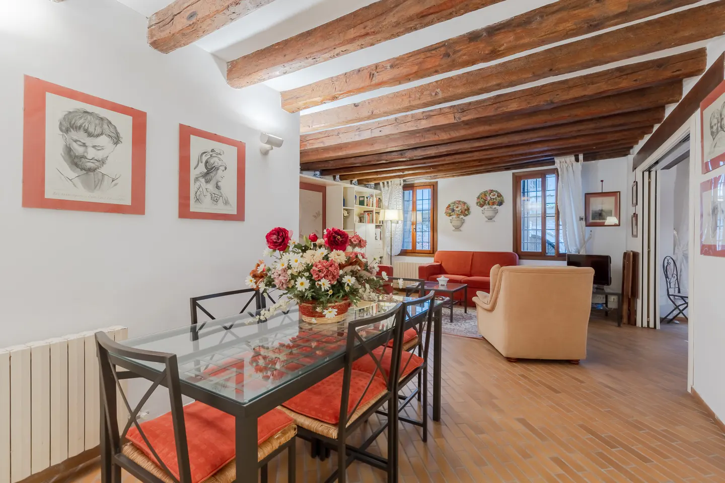 Interior view of a dining and living room with exposed wood beams. A glass table with red cushions sits near two framed portraits. A red sofa and beige chair are in the background.