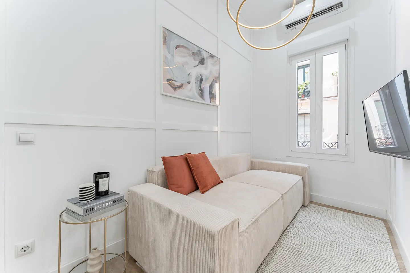 Bright living room with a beige corduroy sofa, rust pillows, and a gold side table with books. A modern light fixture hangs above.