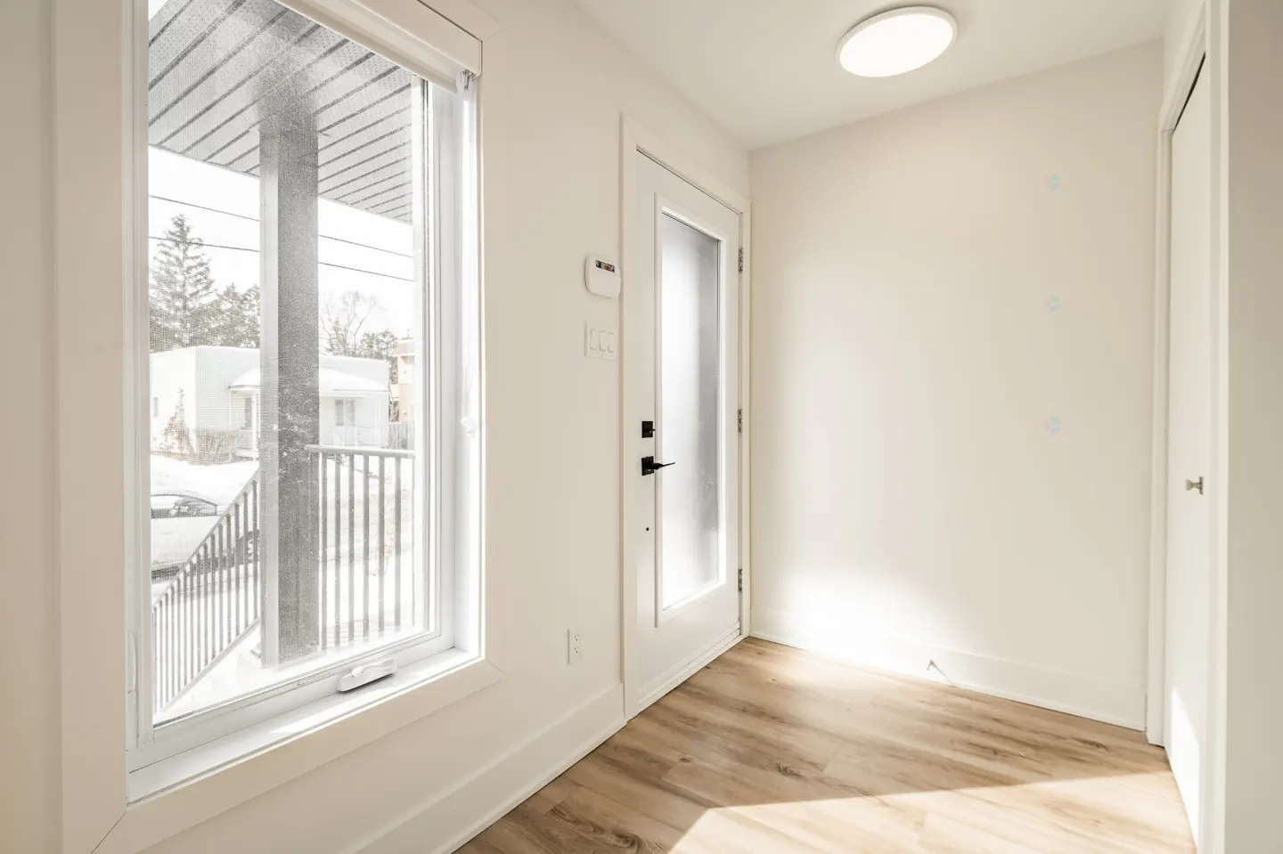 Bright entryway with wood-look floors, white walls, and a window showing a snowy exterior. A white door with a frosted glass pane is visible.
