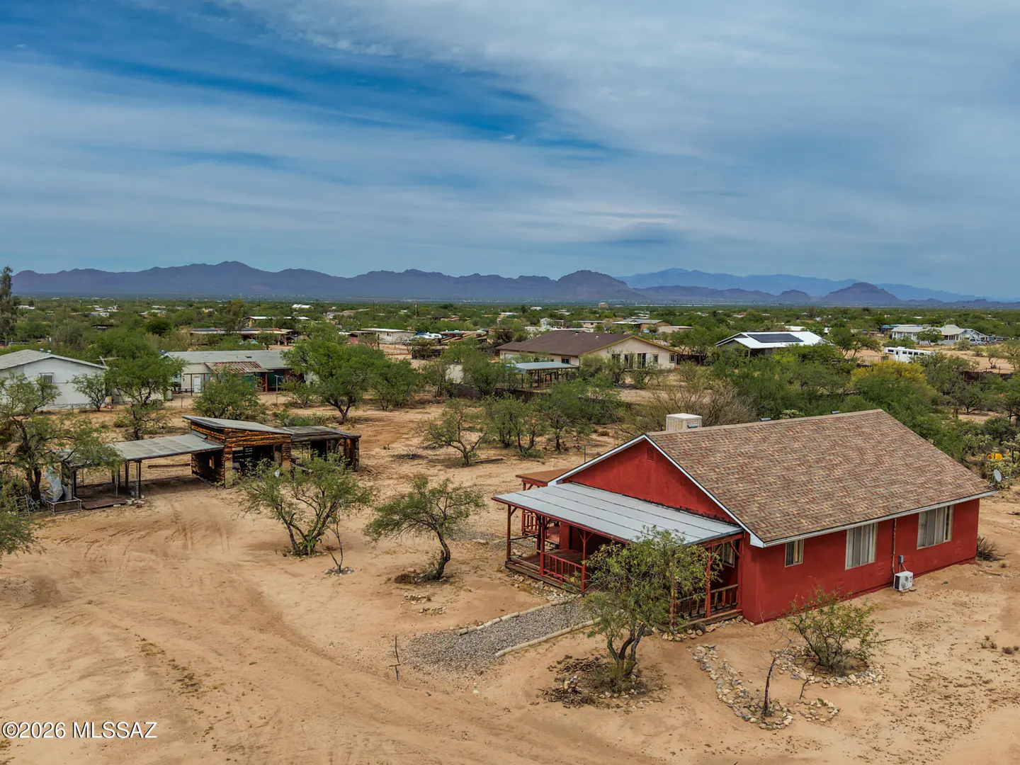 Aerial view of a red house with a porch in a desert landscape, mountains in the distance under a blue sky.