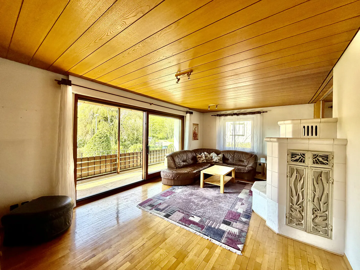 Bright living room with wood floors, ceiling, and a brown leather sectional sofa. A large sliding glass door leads to a balcony.
