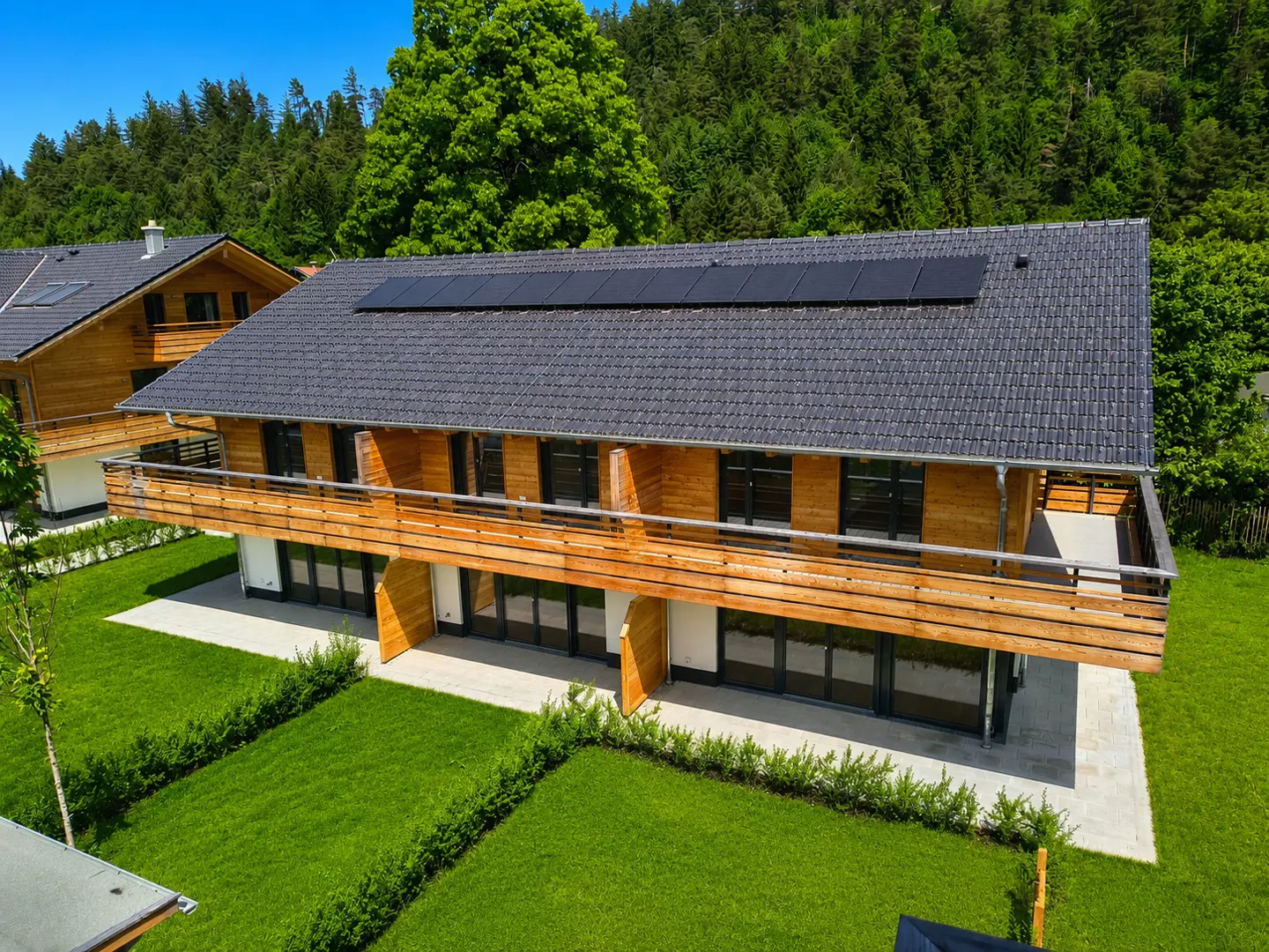 Two-story wood home with a gray tile roof and solar panels. A wooden balcony spans the second floor. Green lawn and trees surround the house.