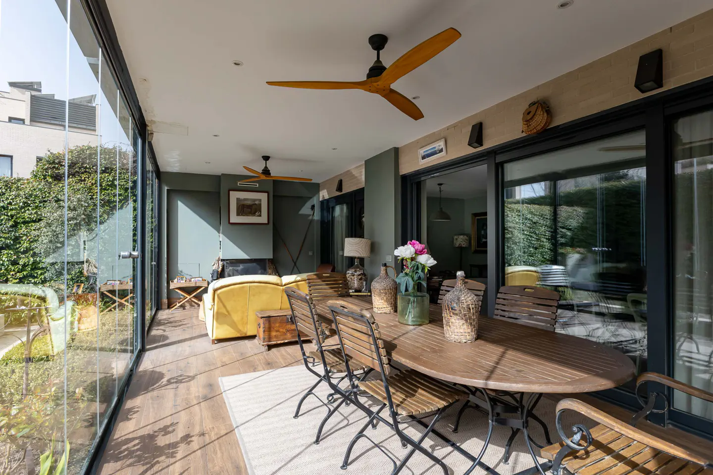 Sunroom with glass walls, wood floors, and ceiling fans. A wooden table and chairs sit on a rug. A yellow couch is visible in the background.